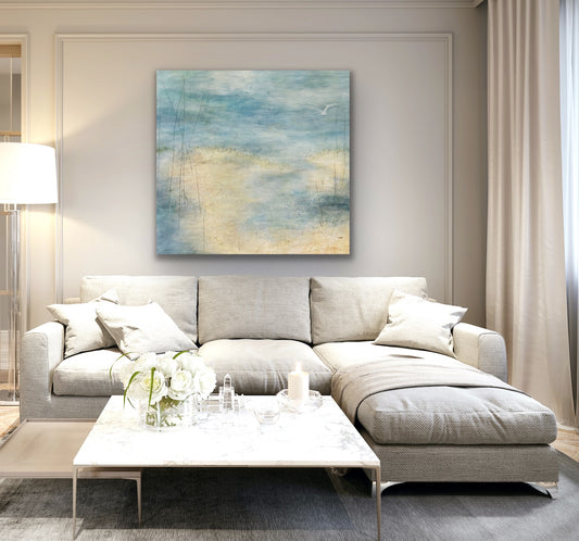 Insitu living room with a white sofa and white walls and an acrylic painting of a soft seascape with water overlapping sand in some areas. There is one lone seagull flying over the water. Painting by Juanita Bellavance, Cumming, GA artist
